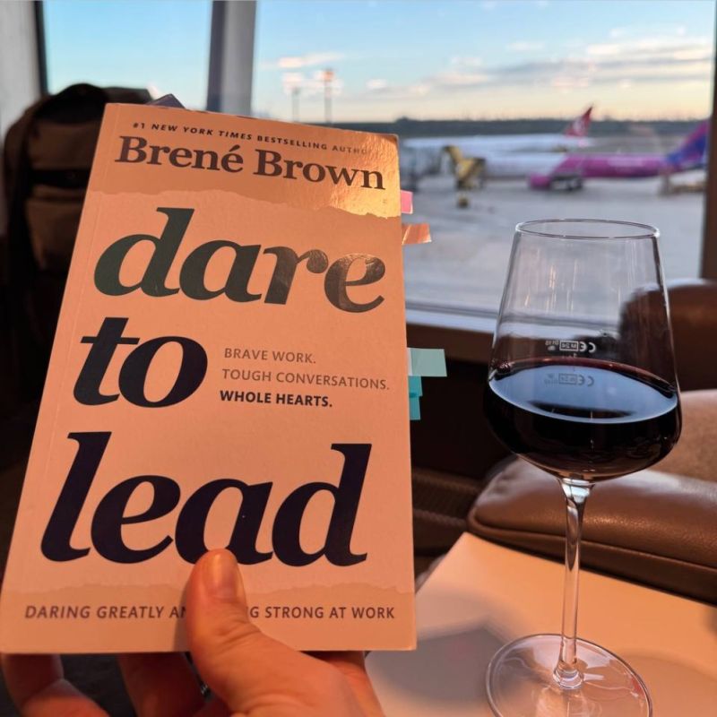 Me holding the book Dare to Lead by Brené Brown in an airport lounge. A glass of red wine is on the table, and airplanes are visible through the large window.